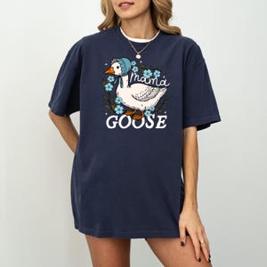 Puede incluir: Una camiseta azul marino con un ganso blanco que lleva un gorro azul y est&aacute; rodeado de flores azules. El texto "mama goose" est&aacute; impreso en la camiseta.