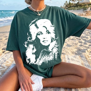 May include: A dark green short-sleeve t-shirt featuring a large, white, high-contrast graphic print of a woman with voluminous hair on the front. The shirt has a crew neckline and a relaxed fit, showcasing a detailed portrait illustration.