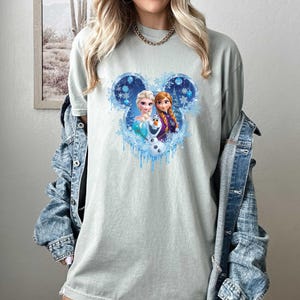 May include: A light green t-shirt featuring a Disney-themed graphic. The graphic showcases Elsa, Anna, and Olaf within a Mickey Mouse head outline, surrounded by snowflakes and icicles. The shirt is a comfortable, casual style.