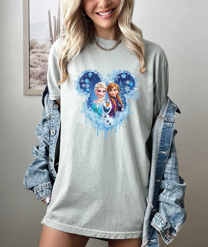May include: A light green t-shirt featuring a Disney-themed graphic. The graphic showcases Elsa, Anna, and Olaf within a Mickey Mouse head outline, surrounded by snowflakes and icicles. The shirt is a comfortable, casual style.