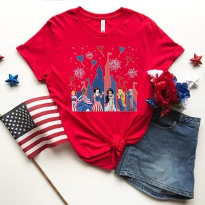 May include: A red t-shirt featuring a patriotic design with cartoon princesses in front of a castle, with fireworks and heart-shaped balloons. The shirt is paired with a denim skirt, decorative stars, and an American flag.