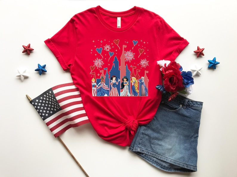 May include: A red t-shirt featuring a patriotic design with cartoon princesses in front of a castle, with fireworks and heart-shaped balloons. The shirt is paired with a denim skirt, decorative stars, and an American flag.