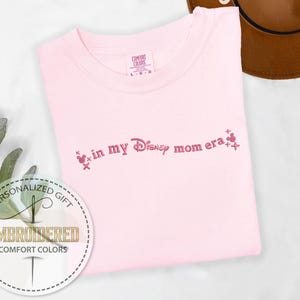 May include: A light pink Comfort Colors t-shirt with the embroidered phrase "in my Disney mom era" in pink script. The design includes Mickey Mouse head icons. A personalized gift.
