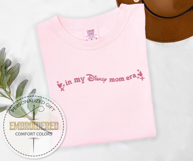 May include: A light pink Comfort Colors t-shirt with the embroidered phrase "in my Disney mom era" in pink script. The design includes Mickey Mouse head icons. A personalized gift.
