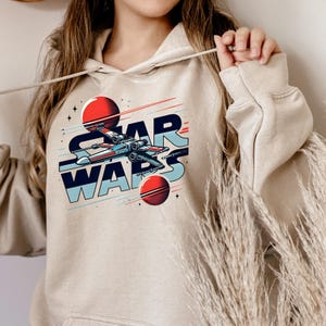 Puede incluir: Una sudadera con capucha beige con un gr&aacute;fico de una nave espacial volando por el espacio con el texto "STAR WARS" en un estilo retro.