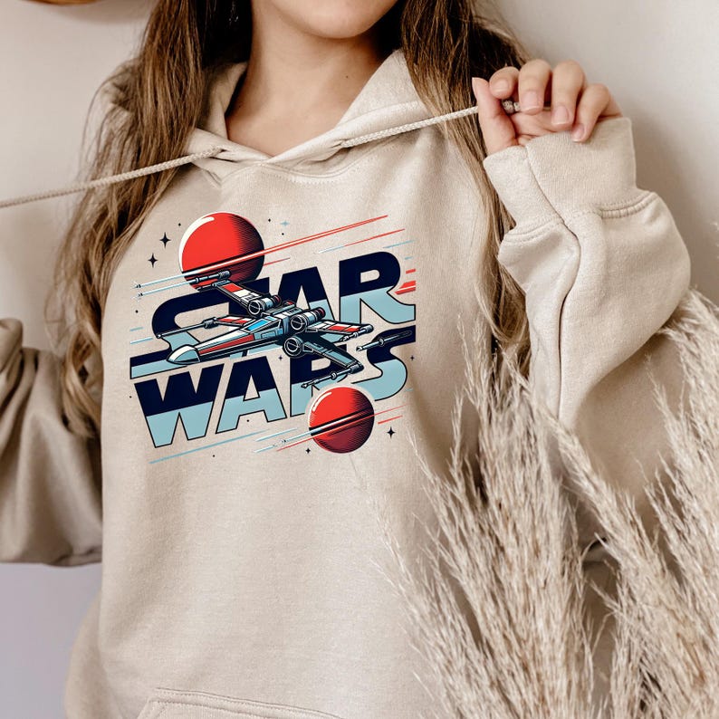 Puede incluir: Una sudadera con capucha beige con un gr&aacute;fico de una nave espacial volando por el espacio con el texto "STAR WARS" en un estilo retro.