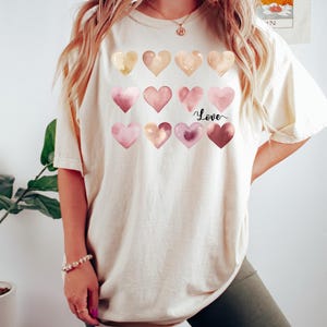 Puede incluir: Una camiseta blanca con un gr&aacute;fico de corazones de acuarela en tonos de rosa y dorado. El texto "Love" est&aacute; escrito en cursiva negra debajo de los corazones.