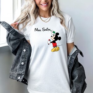 May include: White t-shirt with a Mickey Mouse graphic holding a marker and a paintbrush. The text "Mrs. Smith" is printed above Mickey Mouse.