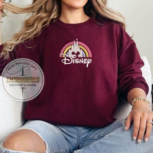 May include: A maroon sweatshirt with a rainbow and castle graphic and the text "Disney" in white.