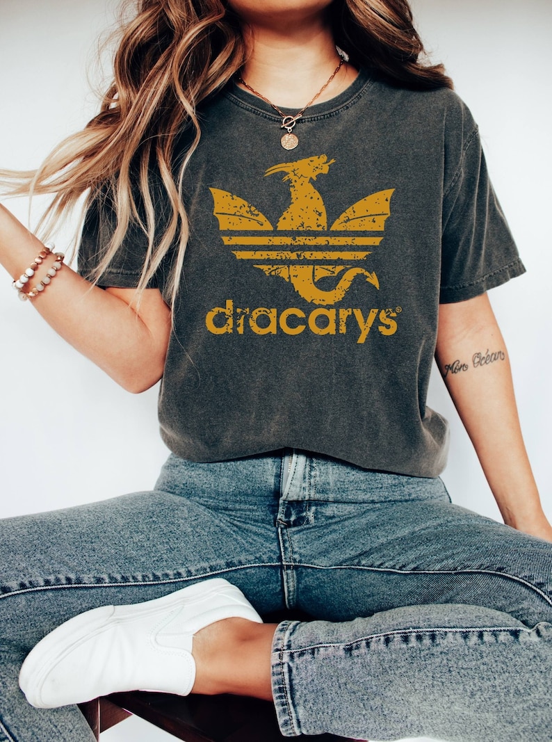 Pu&ograve; includere: Una maglietta grigia scura con un design di drago dorato invecchiato e il testo "dracarys" stampato sul davanti. Il design del drago &egrave; simile al logo Adidas.