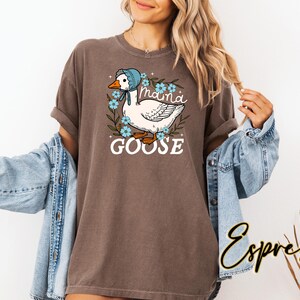 Puede incluir: Una camiseta marr&oacute;n con un ganso blanco que lleva un gorro azul y est&aacute; rodeado de flores azules. El texto "Maina Goose" est&aacute; impreso en la camiseta.