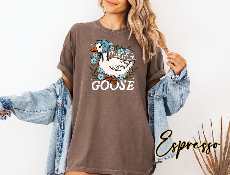 Puede incluir: Una camiseta marr&oacute;n con un ganso blanco que lleva un gorro azul y est&aacute; rodeado de flores azules. El texto "Maina Goose" est&aacute; impreso en la camiseta.
