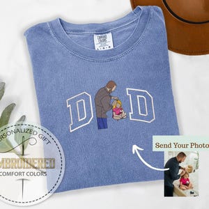 May include: A light blue Comfort Colors t-shirt with the word "DAD" embroidered in white block letters. The "A" is replaced with an illustration of a father and child. The shirt has a crew neck and short sleeves. The words "Personalized Gift" and "Embroidered Comfort Colors" are also visible.