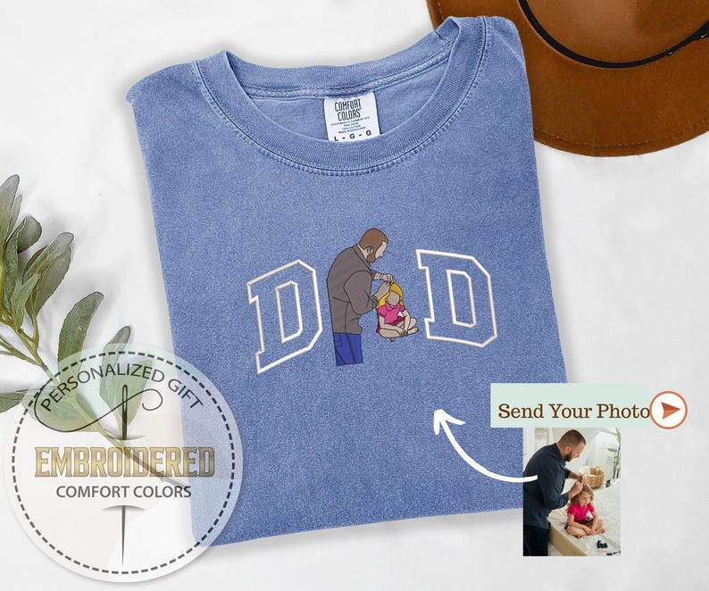 May include: A light blue Comfort Colors t-shirt with the word "DAD" embroidered in white block letters. The "A" is replaced with an illustration of a father and child. The shirt has a crew neck and short sleeves. The words "Personalized Gift" and "Embroidered Comfort Colors" are also visible.