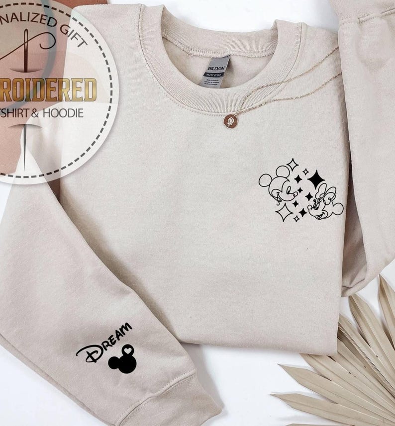 May include: Beige sweatshirt with a black Mickey and Minnie Mouse graphic and the word "DREAM" on the sleeve. A gold necklace with a pendant is draped on the sweatshirt. The sweatshirt is a Gildan brand.
