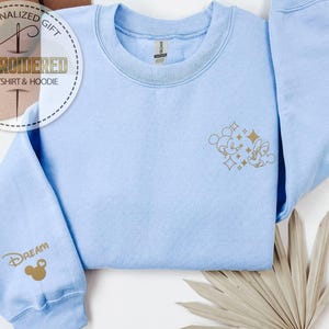 May include: Light blue sweatshirt with gold embroidered Mickey and Minnie Mouse design on the front and the word "Dream" on the sleeve. The sweatshirt has a crew neck and long sleeves. The text "Personalized Gift Embroidered Sweatshirt & Hoodie" is in a circular design.