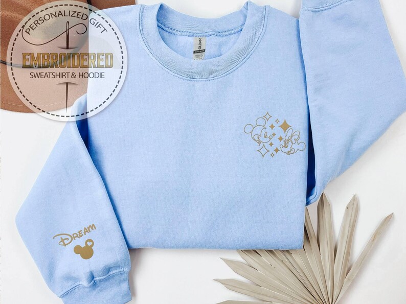 May include: Light blue sweatshirt with gold embroidered Mickey and Minnie Mouse design on the front and the word "Dream" on the sleeve. The sweatshirt has a crew neck and long sleeves. The text "Personalized Gift Embroidered Sweatshirt & Hoodie" is in a circular design.