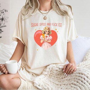 May include: A cream-coloured t-shirt featuring the text "SUGAR SPICE AND FUCK ICE" above a vintage-style illustration of a girl with an ice cream cone inside a red heart. The shirt is worn by a person.