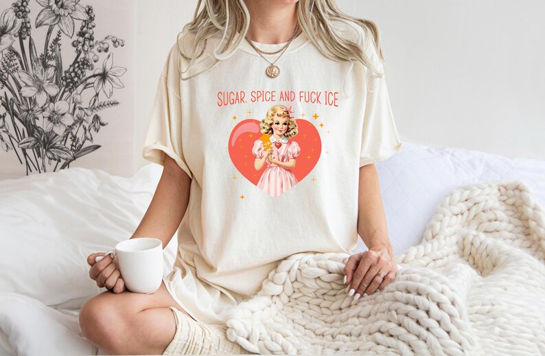 May include: A cream-coloured t-shirt featuring the text "SUGAR SPICE AND FUCK ICE" above a vintage-style illustration of a girl with an ice cream cone inside a red heart. The shirt is worn by a person.