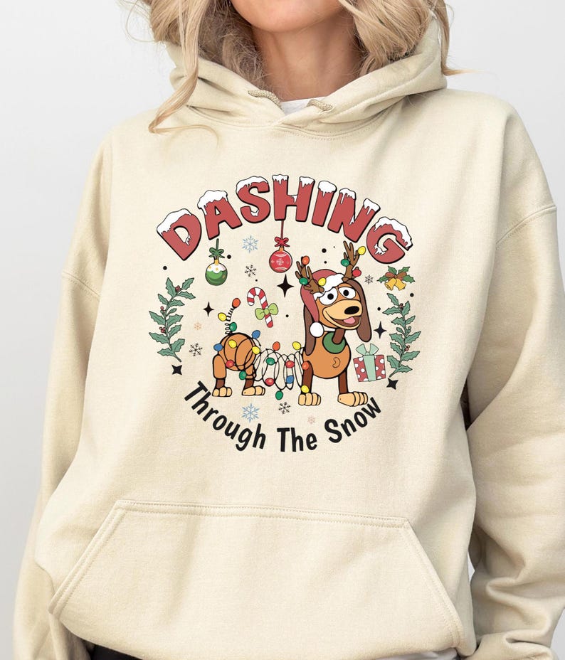 May include: Beige hoodie featuring a festive graphic. The design includes two cartoon dogs adorned with Christmas lights and reindeer antlers, surrounded by holiday ornaments and the text "Dashing Through The Snow."