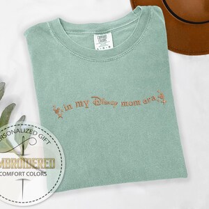 May include: A light green Comfort Colors t-shirt with the embroidered phrase "in my Disney mom era" in a copper color. The shirt has a crew neck and short sleeves. A personalized gift label is in the lower left corner.