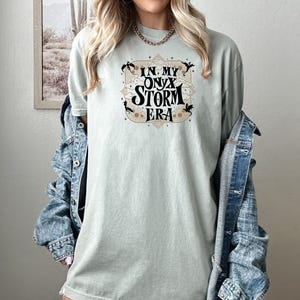 May include: A light green t-shirt with a graphic design that says "In My Onyx Storm Era" in black text with gold accents. The graphic design features a stylized cloud with a dragon on each side.