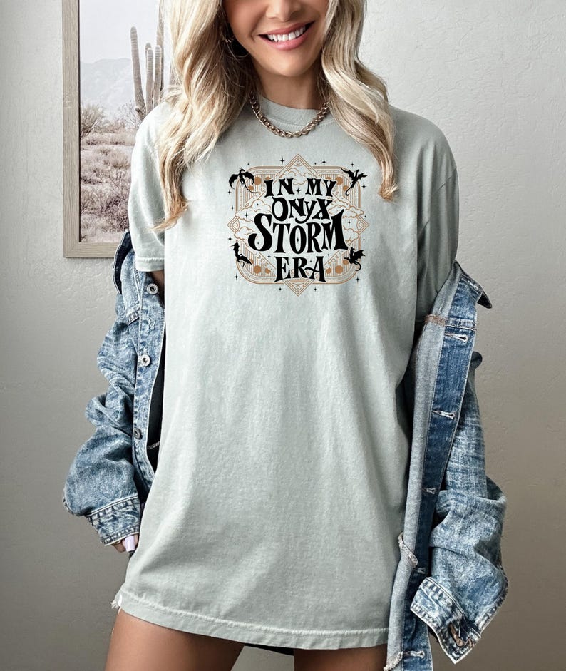 May include: A light green t-shirt with a graphic design that says "In My Onyx Storm Era" in black text with gold accents. The graphic design features a stylized cloud with a dragon on each side.