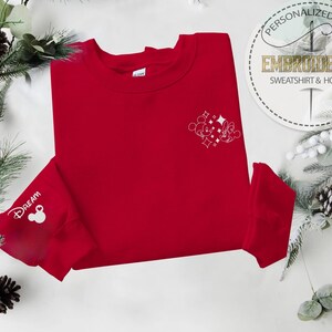 May include: A red sweatshirt with white embroidered Mickey Mouse and Minnie Mouse design on the chest. The left sleeve cuff features the word "Dream" and a Mickey Mouse head. The sweatshirt is surrounded by festive greenery and pinecones.