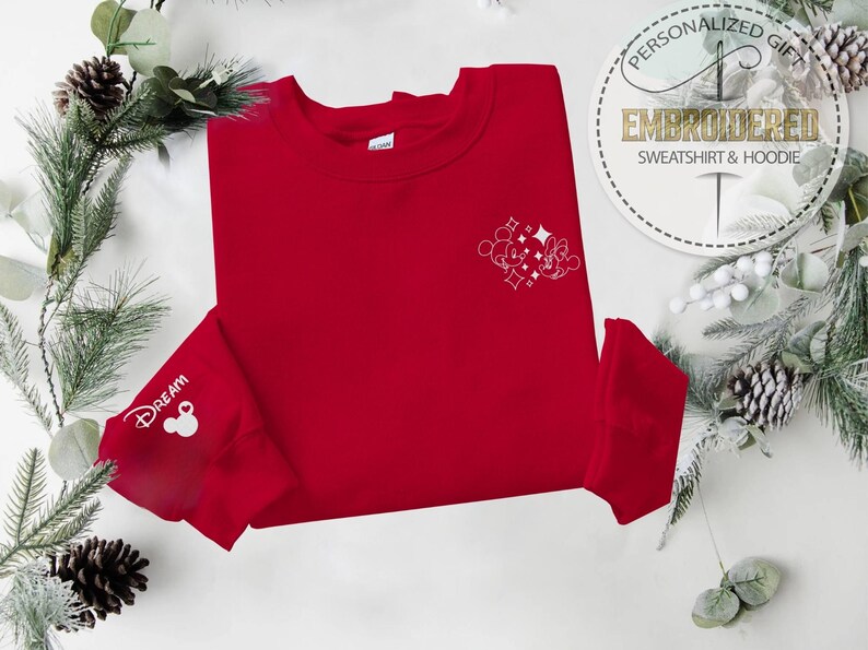 May include: A red sweatshirt with white embroidered Mickey Mouse and Minnie Mouse design on the chest. The left sleeve cuff features the word "Dream" and a Mickey Mouse head. The sweatshirt is surrounded by festive greenery and pinecones.