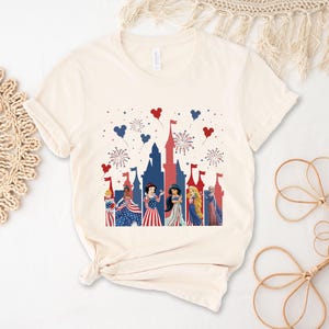 May include: Cream-colored t-shirt featuring a patriotic castle design with fireworks and heart-shaped balloons in red, white, and blue. The graphic includes cartoon characters in festive attire, celebrating a holiday.