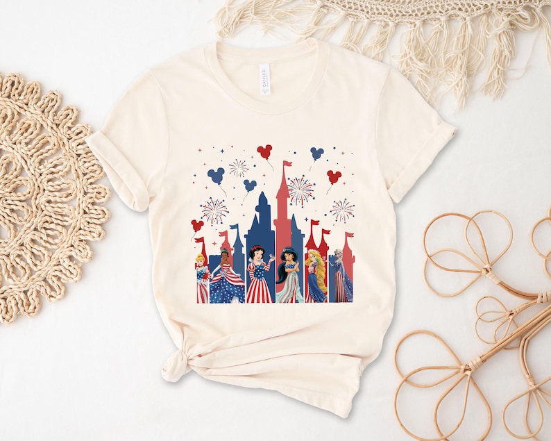 May include: Cream-colored t-shirt featuring a patriotic castle design with fireworks and heart-shaped balloons in red, white, and blue. The graphic includes cartoon characters in festive attire, celebrating a holiday.