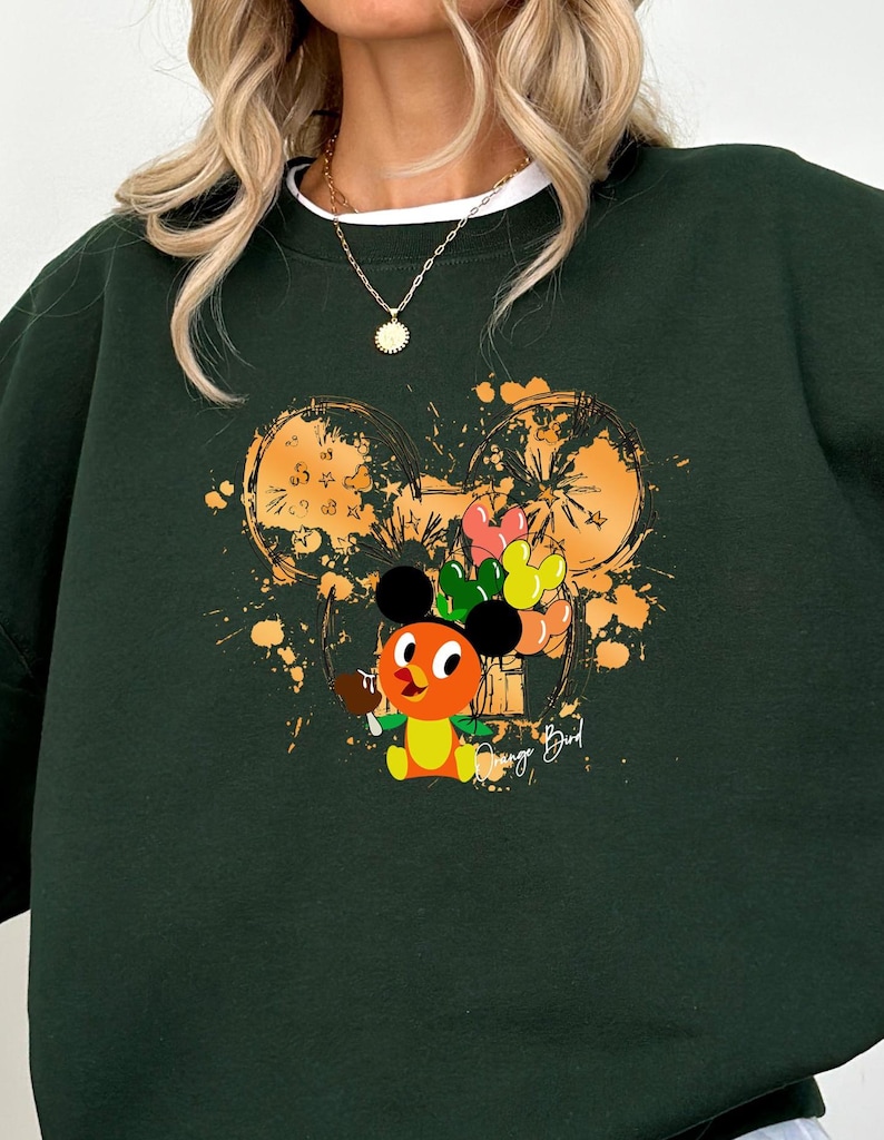 May include: Dark green sweatshirt with a white crew neck collar. The front features a cartoon character holding balloons, with a Mickey Mouse head outline in orange and yellow. The text "Orange Bird" is printed on the right side.