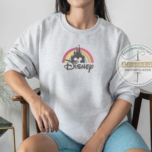 May include: A gray sweatshirt with a rainbow and castle graphic and the text "Disney" in black. The sweatshirt is being worn by a person.