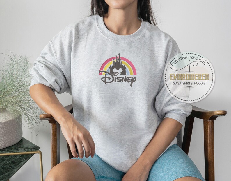 May include: A gray sweatshirt with a rainbow and castle graphic and the text "Disney" in black. The sweatshirt is being worn by a person.