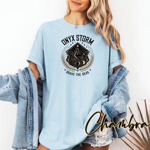 May include: A light blue t-shirt with a graphic design of a black and gold celestial scene. The design features a crescent moon, stars, and a stylized figure. The text "ONYX STORM" and "BRAVE THE DARK" are printed on the shirt.