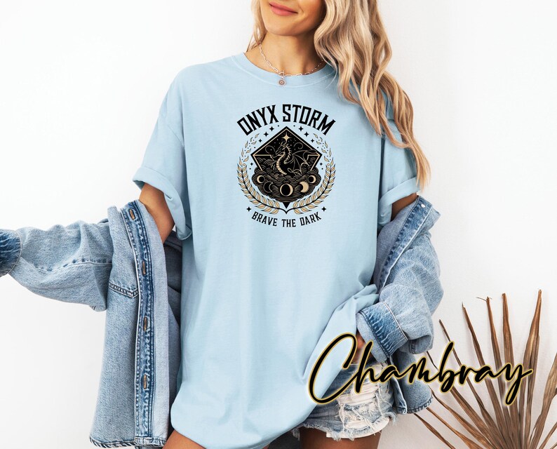 May include: A light blue t-shirt with a graphic design of a black and gold celestial scene. The design features a crescent moon, stars, and a stylized figure. The text "ONYX STORM" and "BRAVE THE DARK" are printed on the shirt.