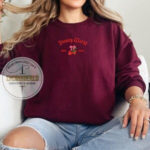 May include: Burgundy crewneck sweatshirt with the text "Disney World" in red script, along with a cherry graphic. The sweatshirt is embroidered and has long sleeves. The text "EST. 1971" is also included.