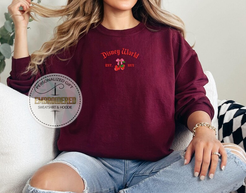 May include: Burgundy crewneck sweatshirt with the text "Disney World" in red script, along with a cherry graphic. The sweatshirt is embroidered and has long sleeves. The text "EST. 1971" is also included.