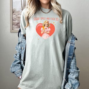 May include: A pale green t-shirt featuring the text "SUGAR, SPICE AND FUCK ICE" above a graphic of a woman holding an ice cream treat inside a heart. The woman is wearing a pink dress. The shirt is a casual style.