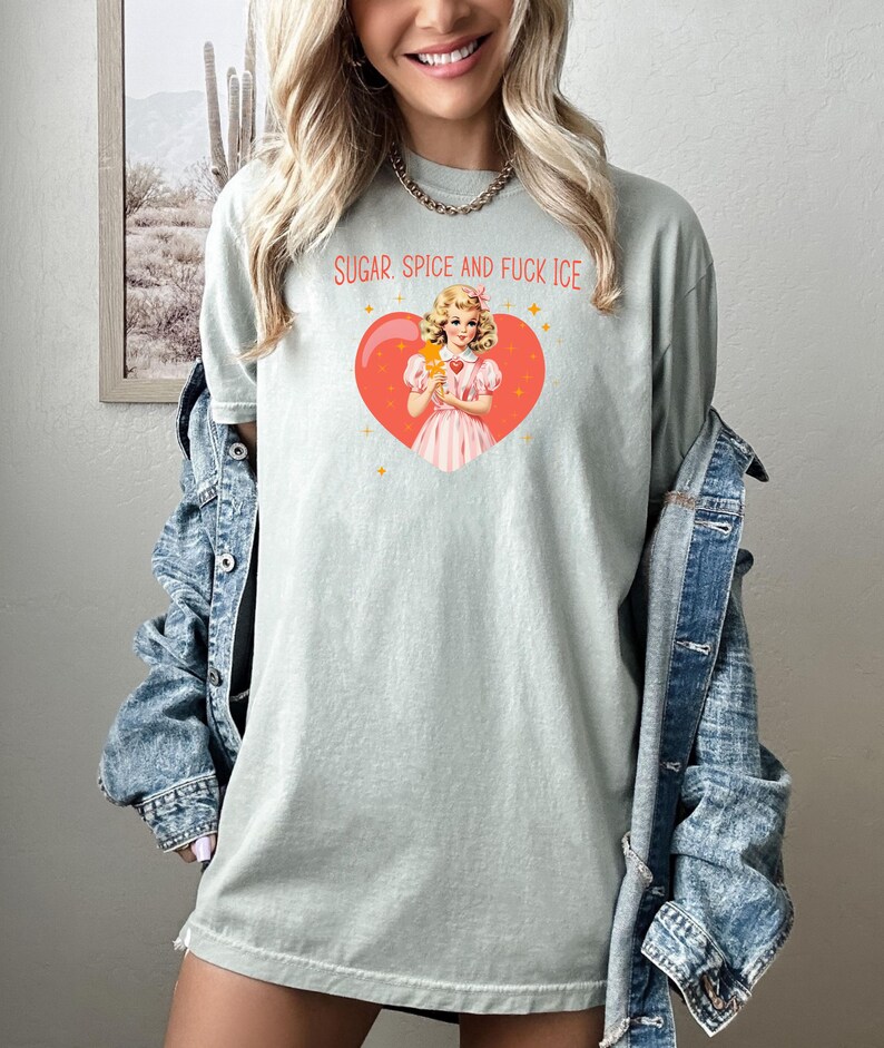 May include: A pale green t-shirt featuring the text "SUGAR, SPICE AND FUCK ICE" above a graphic of a woman holding an ice cream treat inside a heart. The woman is wearing a pink dress. The shirt is a casual style.