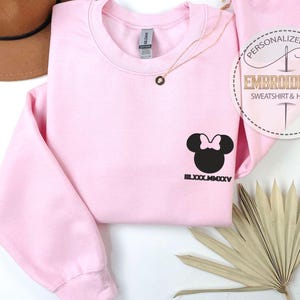 May include: A pale pink sweatshirt featuring a black Minnie Mouse design and the Roman numeral date LXXX.MMXXV. A gold necklace and a decorative palm leaf are also visible. The sweatshirt has the text "PERSONALIZED GIFT EMBROIDERED SWEATSHIRT & HOODIE".