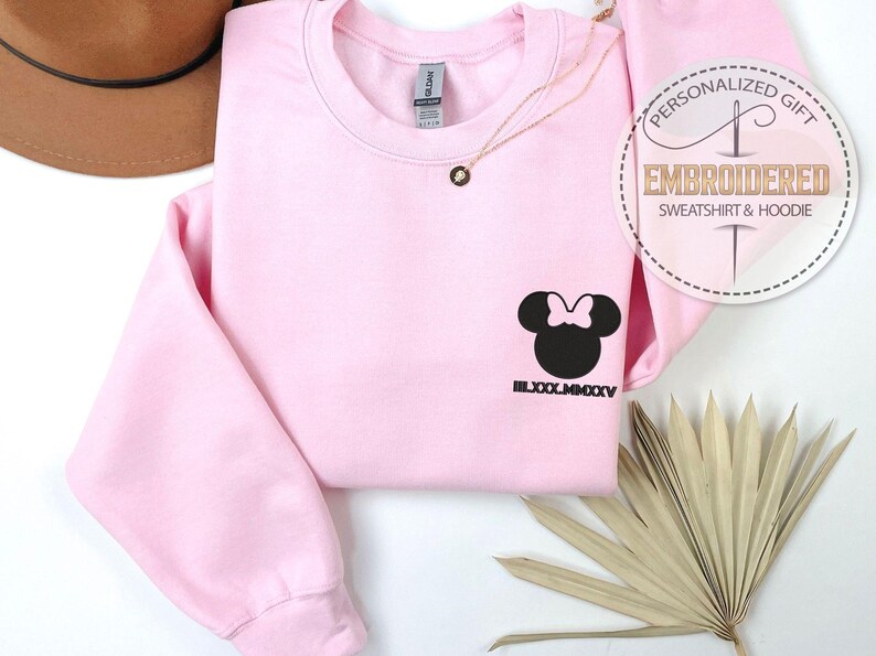May include: A pale pink sweatshirt featuring a black Minnie Mouse design and the Roman numeral date LXXX.MMXXV. A gold necklace and a decorative palm leaf are also visible. The sweatshirt has the text "PERSONALIZED GIFT EMBROIDERED SWEATSHIRT & HOODIE".