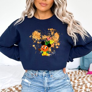 May include: Navy blue sweatshirt featuring a cartoon character with a Mickey Mouse-inspired head, holding balloons. The design includes a gold-colored Mickey Mouse head outline with paint splatters. The sweatshirt is a comfortable, casual style.