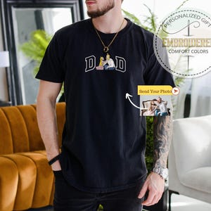 May include: Black t-shirt with the word "DAD" in white block letters and a small image of a couple. The shirt has a round neck and short sleeves. The image also includes a "Send Your Photo" button.