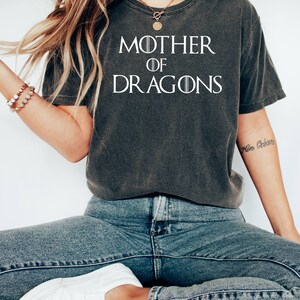 Op de afbeelding: Een donkergrijs T-shirt met de tekst "Mother of Dragons" in wit. Het shirt wordt gedragen door een persoon met lang blond haar en een gouden ketting.