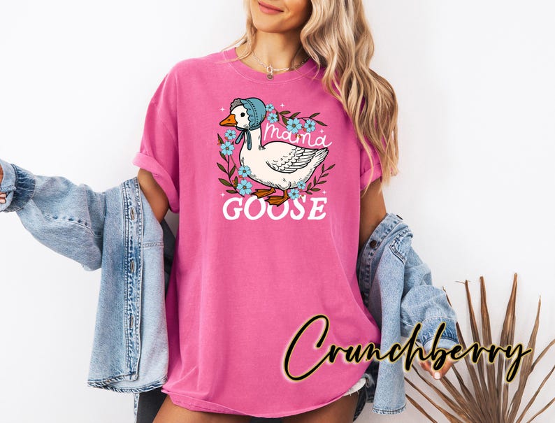 Puede incluir: Camiseta rosa con un ganso de dibujos animados que lleva un gorro azul y el texto "Mama Goose" en una corona de flores.