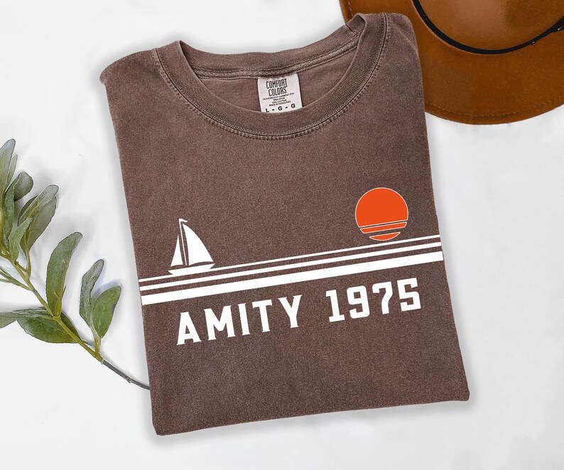 May include: A brown t-shirt with the text "AMITY 1975" in white, along with a sailboat and a sunset graphic. The shirt is folded and has a tag that says "COMFORT COLORS". A brown hat is visible in the background.