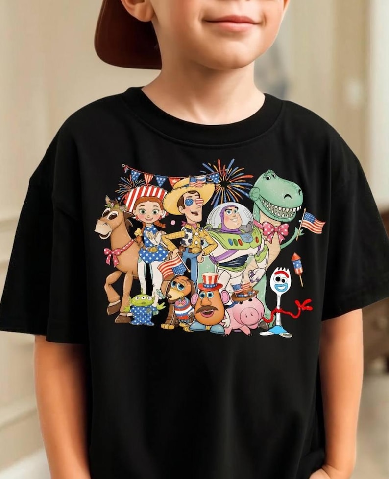 Puede incluir: Una camiseta negra de manga corta con un estampado gr&aacute;fico colorido de varios personajes de juguetes animados. Los personajes est&aacute;n decorados con patrones de banderas, estrellas y rayas, acompa&ntilde;ados de fuegos artificiales y banderines.