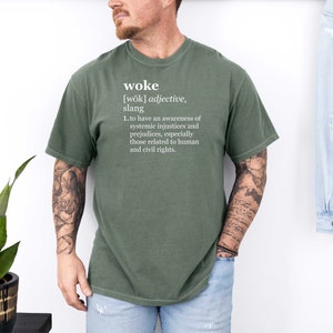 May include: A green t-shirt with the word "woke" defined in white text. The definition reads: "[wōk] adjective, slang 1. to have an awareness of systemic injustices and prejudices, especially those related to human and civil rights."