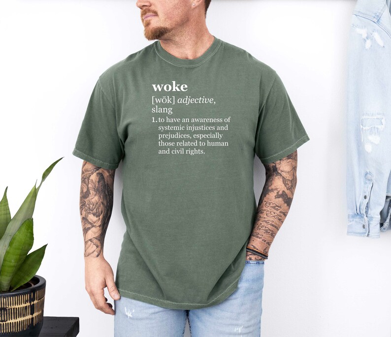 May include: A green t-shirt with the word "woke" defined in white text. The definition reads: "[wōk] adjective, slang 1. to have an awareness of systemic injustices and prejudices, especially those related to human and civil rights."